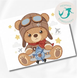 Op de afbeelding: Borduurwerk van een bruine teddybeer met een bruine vliegeniersmuts en een bril, die een blauw en wit vliegtuig vasthoudt. De beer is omringd door witte wolken en gele sterren.