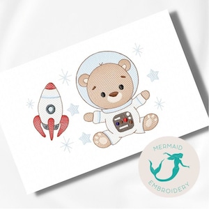 May include: Embroidered design of a brown bear wearing a white astronaut suit with a blue helmet, holding a red and white rocket. The design is surrounded by small white stars.