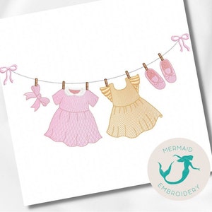 Pode incluir: Dois vestidos de bebê bordados pendurados em um varal com laços e sapatos rosa. Os vestidos são rosa e amarelo com detalhes em branco. O texto "Mermaid Embroidery" está no canto inferior direito.