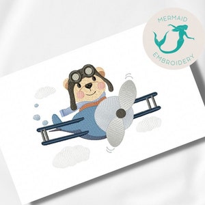Pode incluir: Desenho bordado de um urso de peluche castanho a usar óculos de aviador e um cachecol, a pilotar um avião azul com uma hélice branca. O desenho inclui nuvens brancas.