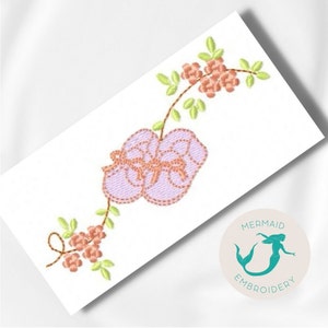 Può includere: Un ricamo che rappresenta un paio di scarpine da bebè rosa con un fiocco, circondato da un tralcio con foglie verdi e fiori rosa. Il disegno è su uno sfondo bianco. L'immagine include anche un logo per Mermaid Embroidery.