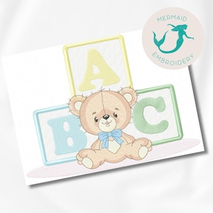 Pode incluir: Um bordado com um urso de pelúcia com uma gravata borboleta azul, em frente a três blocos de alfabeto. Os blocos são azul claro, amarelo e verde, com as letras A, B e C.