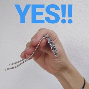 Pu&ograve; includere: Un fermaglio per capelli in argento con un design decorativo. Il fermaglio &egrave; tenuto in una mano con un braccialetto di perline blu e marroni al polso. La parola "YES!" &egrave; scritta in lettere blu sopra la mano.