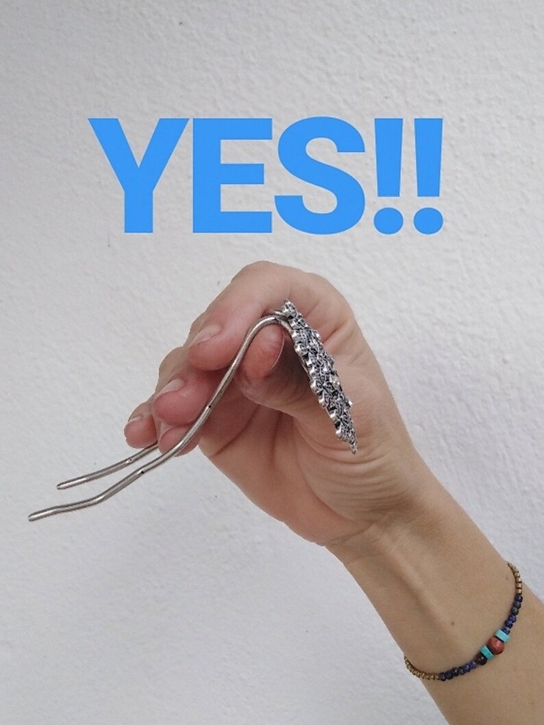 Pu&ograve; includere: Un fermaglio per capelli in argento con un design decorativo. Il fermaglio &egrave; tenuto in una mano con un braccialetto di perline blu e marroni al polso. La parola "YES!" &egrave; scritta in lettere blu sopra la mano.