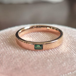 Puede incluir: Un anillo de banda de oro rosa con una pequeña piedra preciosa rectangular verde esmeralda engastada en el centro.