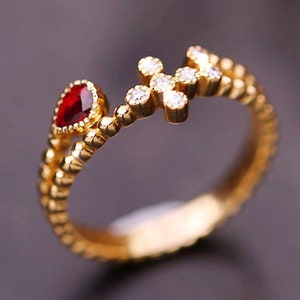 Op de afbeelding: Gouden ring met een rode edelsteen en een klein kruis met heldere stenen. De ring heeft een geperld bandje.