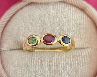 Op de afbeelding: Een gouden ring met drie ovale edelstenen. De edelstenen zijn groen, rood en blauw.