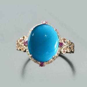 May include: A gold ring featuring a large, oval, bright blue gemstone. The gemstone is surrounded by a halo of small diamonds and accented with pink gemstones. The ring has ornate gold filigree detailing on the band.