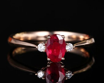 Könnte beinhalten: Ein goldener Ring mit einem großen ovalen Rubin-Mittelstein und zwei kleineren Diamanten auf jeder Seite. Der Ring ist vor einem schwarzen Hintergrund platziert, was einen auffälligen Kontrast erzeugt. Der Rubin hat eine tiefe rote Farbe, und die Diamanten funkeln.