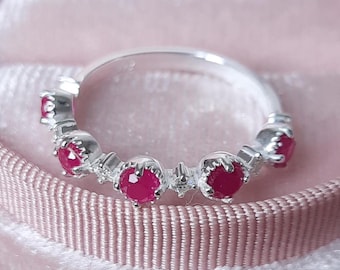 Puede incluir: Un anillo de banda de plata con cinco piedras preciosas rojas engastadas en fila. El anillo está en una caja de terciopelo rosa.