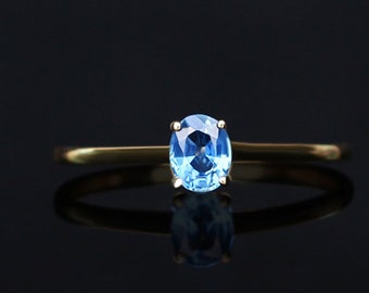 Puede incluir: Un anillo de oro con una piedra preciosa ovalada de color azul claro. El anillo tiene una fina banda de oro, y la piedra preciosa está sujeta por cuatro garras. El anillo se muestra sobre una superficie negra reflectante.