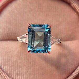 May include: A ring with a large, square-shaped, blue topaz gemstone surrounded by two smaller, triangular-shaped, clear gemstones.