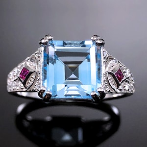 May include: A silver ring with a large, square, light blue gemstone surrounded by smaller diamonds. The ring has two small, red gemstones on either side of the main stone.