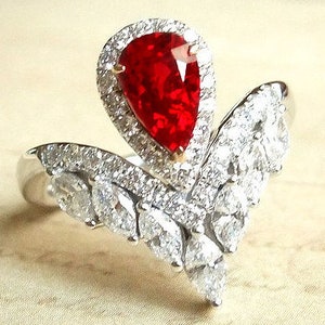 May include: A pear-shaped ruby gemstone ring set in a white gold band. The ring features a crown-like design with a halo of smaller diamonds surrounding the ruby. The band is adorned with additional diamonds in a marquise shape.