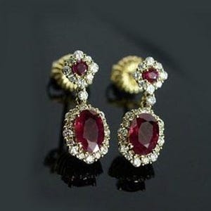 May include: A pair of gold earrings with a ruby and diamond design. Each earring features a large oval ruby surrounded by a halo of small diamonds. The top of each earring has a smaller ruby and diamond cluster.