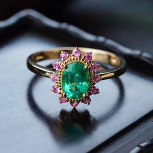 Puede incluir: Un anillo de oro con una piedra central ovalada de esmeralda, rodeada de piedras rosas. El anillo tiene una banda de oro lisa, y las piedras están engastadas en un diseño de halo. El anillo se muestra sobre una superficie oscura.