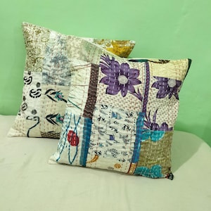 Pode incluir: Duas almofadas decorativas com designs de patchwork. Uma almofada apresenta um grande padrão floral roxo, enquanto a outra tem uma mistura de padrões em bege, verde e branco. As almofadas estão em uma superfície clara.