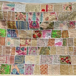 May include: A patchwork textile with a variety of patterned fabric squares in beige, pink, green, and blue. The squares are stitched together with white thread, creating a textured surface. The textile is likely a decorative item, such as a wall hanging or throw.
