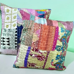 Op de afbeelding: Twee decoratieve sierkussens met een patchwork ontwerp. De kussens hebben een verscheidenheid aan stoffen in tinten roze, paars, groen en oranje. Het patchwork is aan elkaar gestikt met witte draad, wat een textuur creëert.