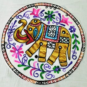 Könnte beinhalten: Runder, gestickter Elefant in Gold, Rot, Blau, Rosa und Grün auf weißem Hintergrund. Der Elefant ist mit komplizierten Mustern verziert und von floralen und Rankenmotiven umgeben. Der Rand ist schwarz mit einem roten und braunen Innenrand.