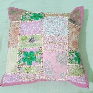 May include: Square decorative pillow with a patchwork design. The pillow features a variety of pink, green, and cream-colored fabrics, with embroidered floral and paisley patterns. The edges are trimmed with a pink border.