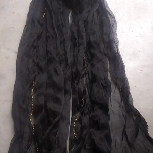 May include: A flowing black scarf with a delicate, sheer texture. The edges are adorned with a line of small, golden beads, adding a touch of elegance. The scarf is draped, showcasing its length and the subtle sheen of the fabric.
