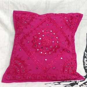 May include: A pink square throw pillow with a decorative embroidered design featuring a central motif of mirrored circles surrounded by floral patterns.