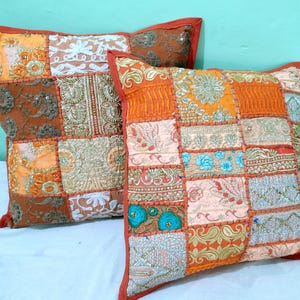 Könnte beinhalten: Zwei dekorative Zierkissen mit Patchwork-Designs. Die Kissen weisen eine Vielzahl von Stoffen in Orange-, Braun- und Cremetönen mit aufwendigen Stickereien und Verzierungen auf. Die Kissen haben einen roten Rand und liegen auf einer weißen Oberfläche.