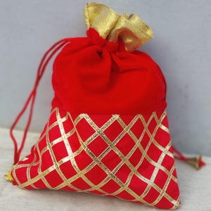 May include: Red velvet drawstring bag with a gold patterned bottom. The bag is closed with a drawstring and has a gold tassel.