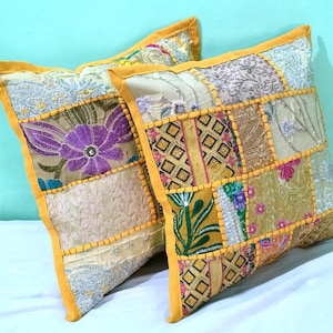 May include: Two decorative cushions with a patchwork design. The cushions feature a variety of fabrics in shades of gold, beige, and purple, with floral and geometric patterns. The edges are trimmed with a bright yellow border.