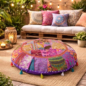 Puede incluir: Un puf de suelo redondo de patchwork en colores vibrantes, incluyendo morado, rosa y dorado. El puf presenta una variedad de patrones de tela y está bordeado con un borde morado y borlas coloridas. Se encuentra sobre una alfombra tejida, con un sofá y cojines en el fondo.