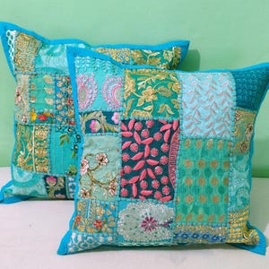 May include: Two square patchwork cushions in teal, turquoise, and gold. The cushions have embroidered floral and geometric patterns. The edges are trimmed with a bright blue border. Each cushion is approximately 40cm x 40cm.