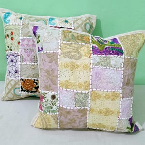 Puede incluir: Dos cojines decorativos de patchwork. Cada cojín presenta cuadrados de tela en tonos pastel, incluyendo crema, lavanda y dorado, con diseños florales y geométricos. Los cojines tienen un borde color crema y están colocados sobre un fondo verde claro.