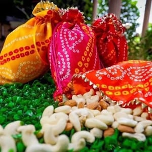 Op de afbeelding: Drie kleurrijke stoffen zakjes met een traditioneel Indiaas ontwerp, gevuld met cashewnoten en amandelen. De zakjes zijn versierd met gouden randjes en liggen op een bed van groen gras.