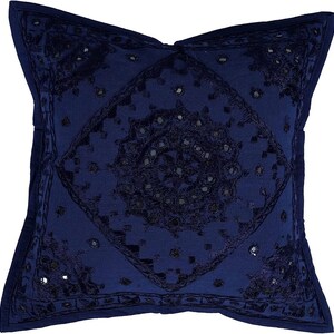 May include: A navy blue cushion with an embroidered design featuring a central motif of mirrored circles. The cushion is square shaped and has a fringed border.