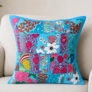 May include: A square throw pillow featuring a vibrant patchwork design in shades of bright blue, magenta, and white. The surface is decorated with intricate floral embroidery, geometric patterns, and stitched fabric panels with a blue border.