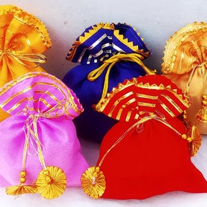 May include: Five small fabric gift bags in different colours. The bags are decorated with gold trim and tassels. The bags are arranged in a row on a white surface.