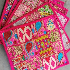 May include: Set of six pink fabric placemats with colourful embroidered patchwork designs. Each placemat has a pink border and features a variety of patterns, including paisley, floral, and geometric designs.
