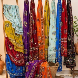 Puede incluir: Una colección de bufandas rectangulares en varios colores, incluyendo morado, rojo, naranja, amarillo, azul y negro. Cada bufanda presenta un bordado intrincado con pequeños espejos circulares y borlas decorativas en los bordes.