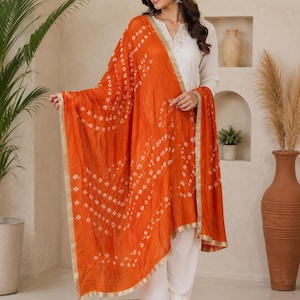 May include: A vibrant orange dupatta featuring a white tie-dye pattern and a metallic gold border. The rectangular fabric is draped over a white long-sleeved top and white trousers, showcasing a traditional textile design with a crinkled texture.