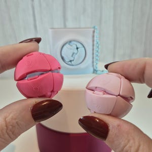 Op de afbeelding: Twee roze, bolvormige puzzelspeelgoed worden tussen de vingers gehouden. Het speelgoed bestaat uit verschillende gebogen segmenten die geopend kunnen worden. Op de achtergrond staat een wit en blauw kubusvormig bakje, samen met een roze bakje.