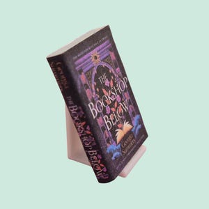 Op de afbeelding: Een hardcover boek getiteld The Bookshop Below door Georgia Summers, met een donkerpaarse en zwarte kaft met een illustratie van een sierlijke boog, bloemmotieven en een afbeelding van een open boek, rechtop op een witte standaard.