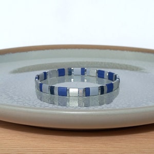Könnte beinhalten: Ein Armband mit einem sich wiederholenden Muster aus blauen, silbernen und hellblauen rechteckigen Perlen. Das Armband wird auf einem hellgrauen Teller mit braunem Rand vor weißem Hintergrund präsentiert.
