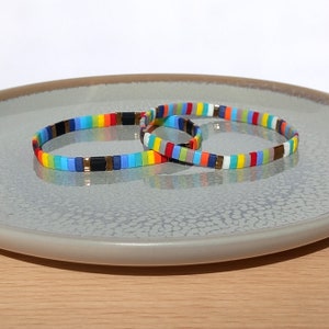 May include: Two colourful beaded bracelets with a rainbow pattern. The bracelets are made of small, square beads in various colours, including red, orange, yellow, green, blue, and purple. The bracelets are displayed on a grey plate.