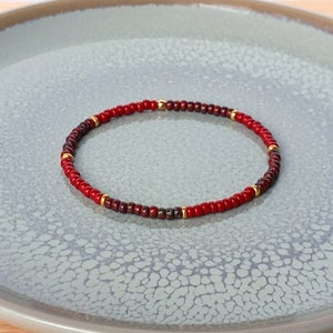 Op de afbeelding: Een rode en bruine kralen armband met gouden accenten. De armband ligt op een grijs, getextureerd oppervlak.