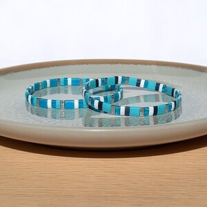 Könnte beinhalten: Zwei blau-weiß gestreifte Armbänder mit silbernen Akzenten. Die Armbänder liegen übereinander auf einem grauen Teller.