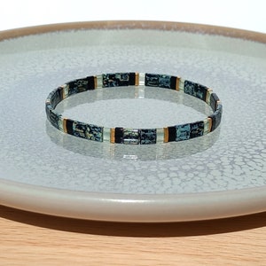May include: A teal, black and gold beaded bracelet with a marbled effect. The bracelet is on a white plate with a textured surface.