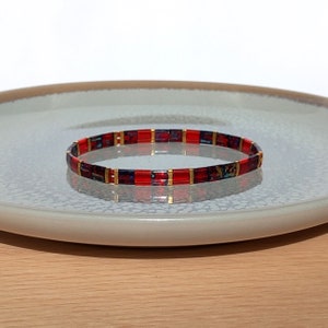 Könnte beinhalten: Ein rotes und goldenes Armband aus kleinen quadratischen Fliesen. Das Armband liegt auf einem grauen Teller.