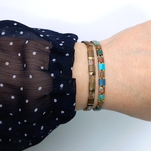 Op de afbeelding: Twee gouden armbanden met kleurrijke vierkante tegels. De ene armband heeft bruine, oranje en blauwe tegels. De andere armband heeft turquoise, blauwe en bruine tegels.