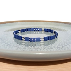 Può includere: Un braccialetto blu e argento fatto di perline quadrate. Il braccialetto è su un piatto bianco.